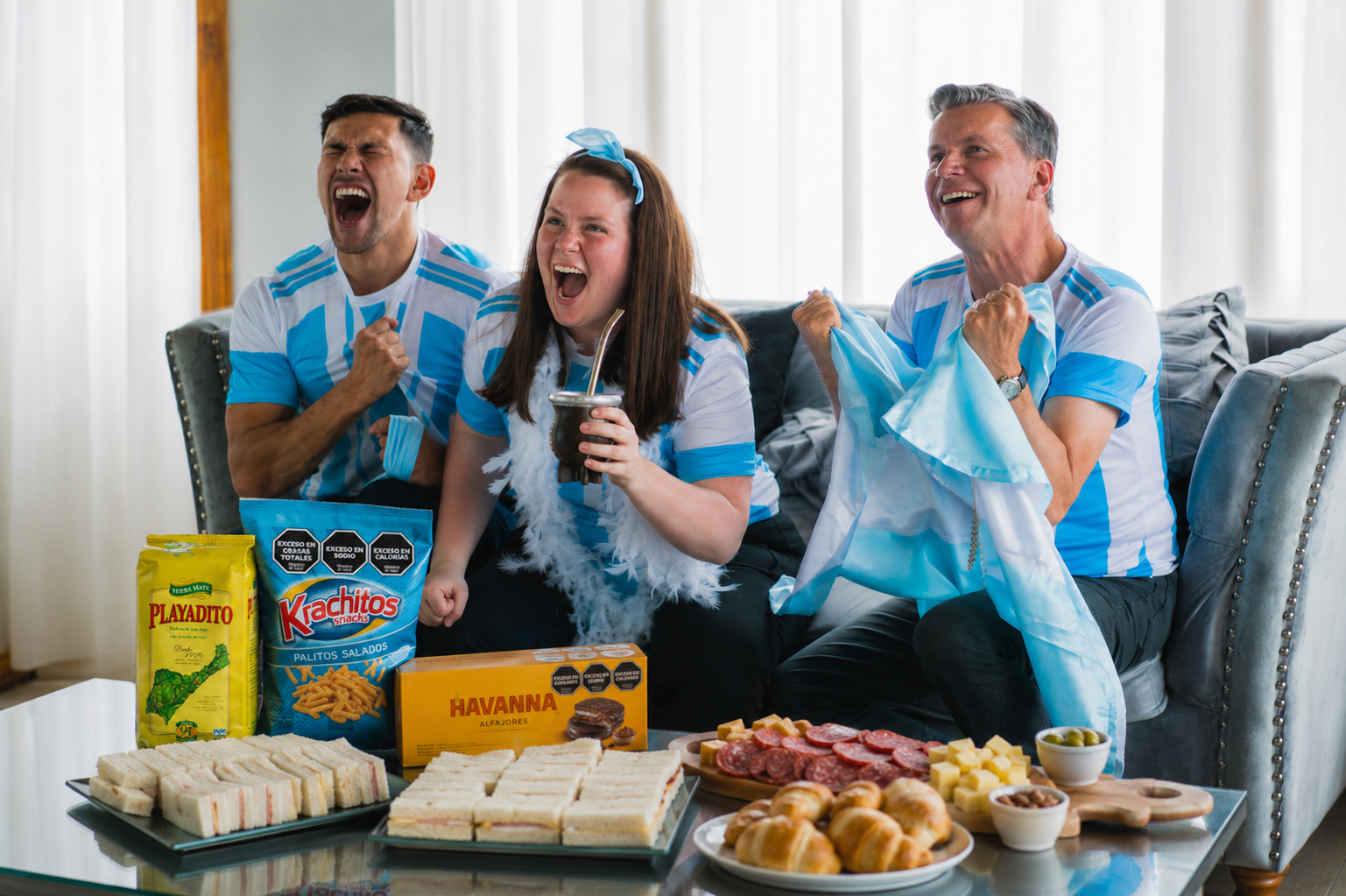 Grupo de personas alentando a la Selecci&oacute;n Argentina durante un partido del Mundial, con camisetas argentinas, mate y productos de Pampa Direct como yerba Playadito, alfajores Havanna, snacks argentinos y comida t&iacute;pica sobre la mesa.