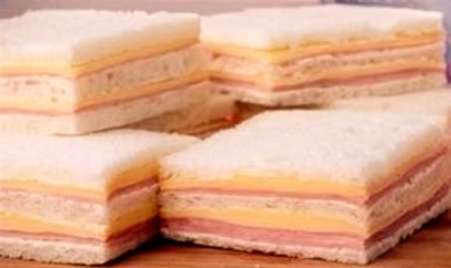 Sándwiches de miga cuadrados apilados, con capas de pan blanco sin corteza, jamón y queso sobre una tabla. Conseguí pan de miga para hacer sandwich de miga en pampadirect.com con envíos a todo el mundo.