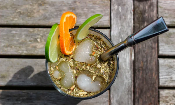 Top view of a tereré glass with ice, lime and orange slices, and a bombilla on a wooden table