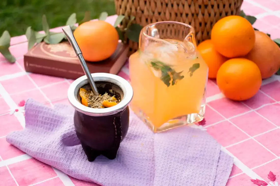 Mate con rodajas de naranja y vaso de jugo con hielo y menta sobre mantel al aire libre
